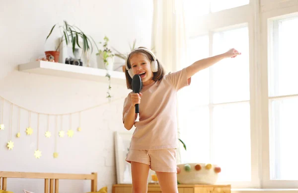 私は超スターです 幸せな就学前の子供は マイクとして毛ブラシを保持し 自宅でベッドの上に立っている間 彼女のお気に入りの歌を歌います かわいいです女の子は有名な歌手であるふり — ストック写真