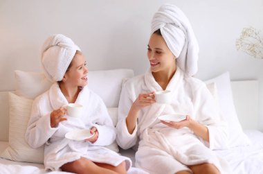 Mutlu, genç bir aile annesi ve kızı sabahları yatakta sıcak bir içecek içerken birbirlerine gülümseyip bakıyorlar.