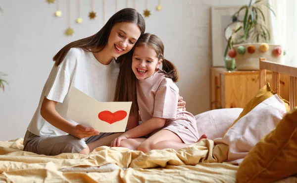 乐观的妇女和女孩坐在床上笑着拥抱 在家里心情愉快地看贺卡 — 图库照片