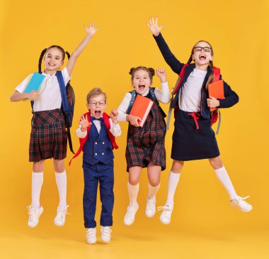 Okul kıyafetleri içinde, sırt çantaları ve kitaplarıyla eğlenen ve sarı arka planda zıplayan bir grup eğlenceli sınıf arkadaşı.