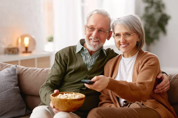 年轻漂亮的老夫妇夫妇一边吃爆米花 一边看电视 同时在客厅的沙发上休息 笑着与成熟的家人拥抱在一起享受周末 休闲活动 — 图库照片