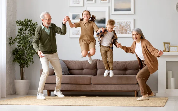 Full length of two excited kids siblings having fun playing with active and positive grandparents in the living room at home, enjoying weekend time with energetic senior old grandma and grandpa