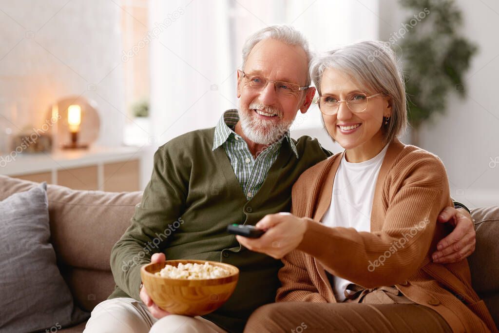 Beautiful happy senior couple husband and wife eating popcorn and watching TV while relaxing on sofa in the living room. Smiling mature family cuddling enjoying weekend together. Leisure activities
