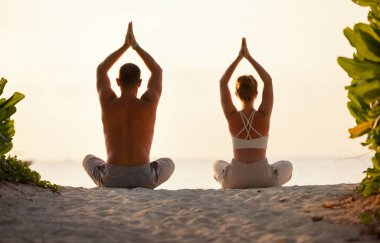 Namaste 'nin elleri kıyı şeridinde başlarının üstündeyken Lotus pozu veren spor elbiseli kadın ve erkeğin arka görüntüsü.