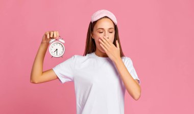 Yastıklı ve çalar saati olan genç bir kadın ağzını kapatıyor ve pembe arka plana karşı sabahın erken saatlerinde uyandıktan sonra gözleri kapalı esniyor.