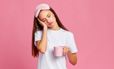 Sabahın köründe pembe arka planda kahve içerken yüzüne dokunan uykulu, genç bir kadın.