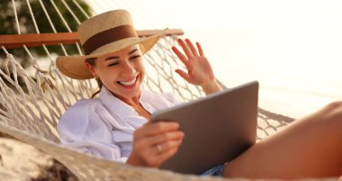 Çevrimiçi konuşma. Hasır şapka takan ve gülümseyen genç, mutlu bir kadın dijital tablette video çağrısı yaparken güneşli bir günde hamakta dinleniyor.