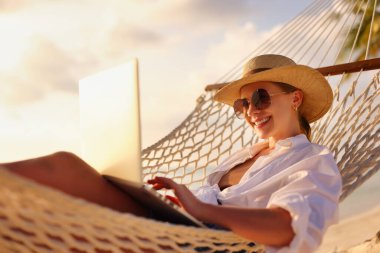 Uzaktan çalışıyor. Genç mutlu kadın, hasır şapkalı ve güneş gözlüklü kadın güneş gözlüklü, gün batımında plajda hamakta dinlenirken. Tatil sırasında uzaklık işi
