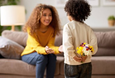 Küçük oğlan, annesinin arkasında çiçek buketi saklarken annesi de Afro-Amerikan yarış kadınını heyecanla koltukta oturmuş bekliyor, seçici bir şekilde. Anneler Günü kutlaması