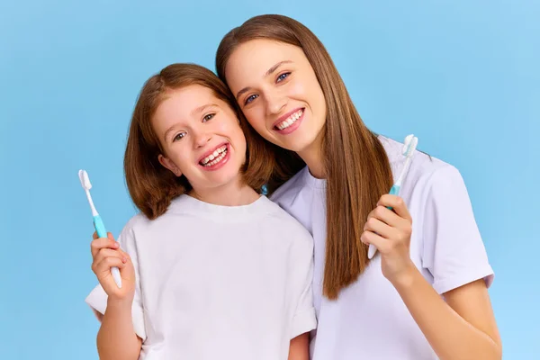 乐观的家庭 身穿白色T恤的妇女和女孩在蓝色背景下一起刷牙时微笑着看着相机 — 图库照片