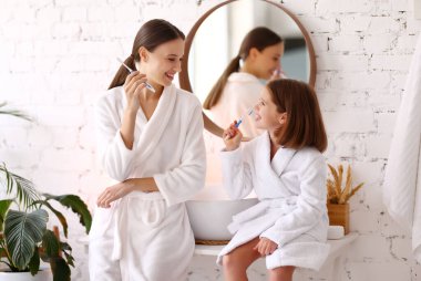 Beyaz bornozlu küçük kızı olan mutlu genç bir kadının banyoda dişlerini fırçalamasının ve birlikte sabah vaktinin tadını çıkarmasının yan görüntüsü.