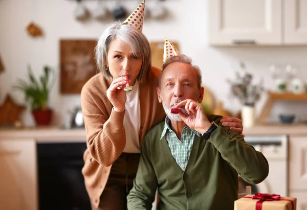 家で誕生日を祝う幸せな美しいシニアカップル パーティー帽子をかぶっている高齢者の男性と女性 笛を吹いてカメラを見てください お祝いと休日のコンセプト — ストック写真
