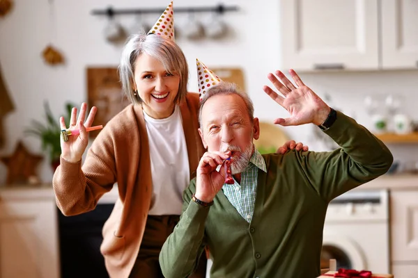 正の高齢者のカップル自宅で誕生日を祝う カメラで手を振る パーティーの帽子で幸せな高齢者の男性と女性が笛と笑顔を吹いて 人々に選択的な焦点 お祝いのコンセプト — ストック写真