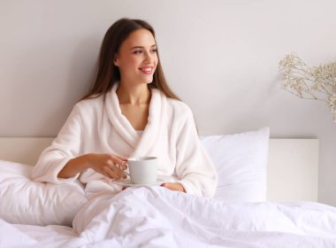 İyimser genç bayan yatakta dinlenirken ve evde sıcak bir içecek içerken gülümsüyor ve başka tarafa bakıyor.