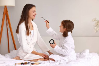 Cilt bakımı sırasında yatakta dinlenirken mutlu bir kızın yanaklarına pudra süren bornozlu neşeli genç bir kadın.