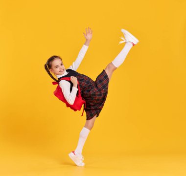 Sırt çantalı, kollarını ve bacaklarını kaldırarak okula koşan, sarı arka planda koşan, kareli elbiseli, gülen ilkokul öğrencisinin tüm vücudu.