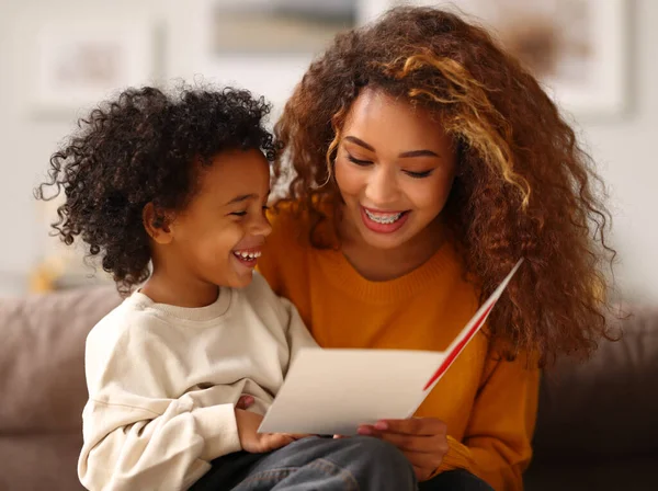Cute little boy son congratulating his mom happy mixed race woman with Mothers day, giving her handmade greeting postcard with red heart while sitting together on sofa at home. Family holidays concept