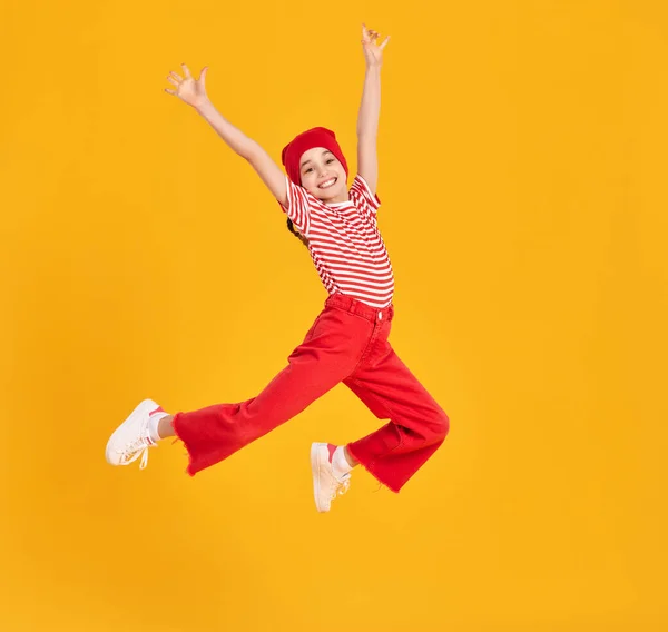 Full body of energetic child girl in striped shirt and red hat and pants raising arms and jumping high against yellow background
