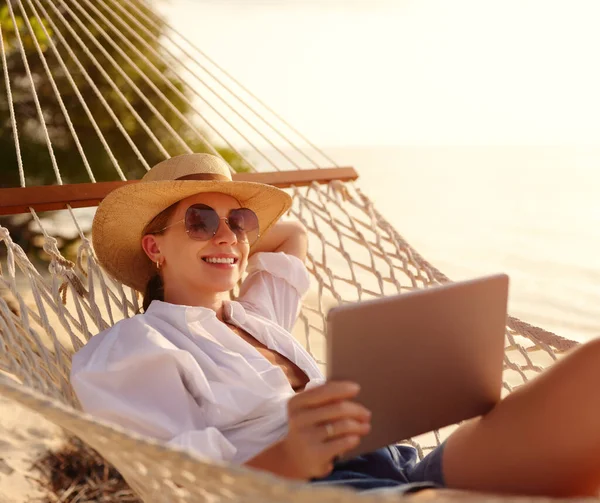 지푸라기 모자를 선글라스를 모래사장의 해먹에서 쉬면서 디지털 태블릿을 사용하며 행복하게 — 스톡 사진