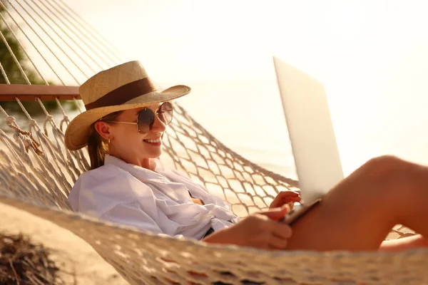 원격으로 합니다 지푸라기 모자를 선글라스를 해변의 해먹에서 휴식을 취하며 노트북으로 — 스톡 사진