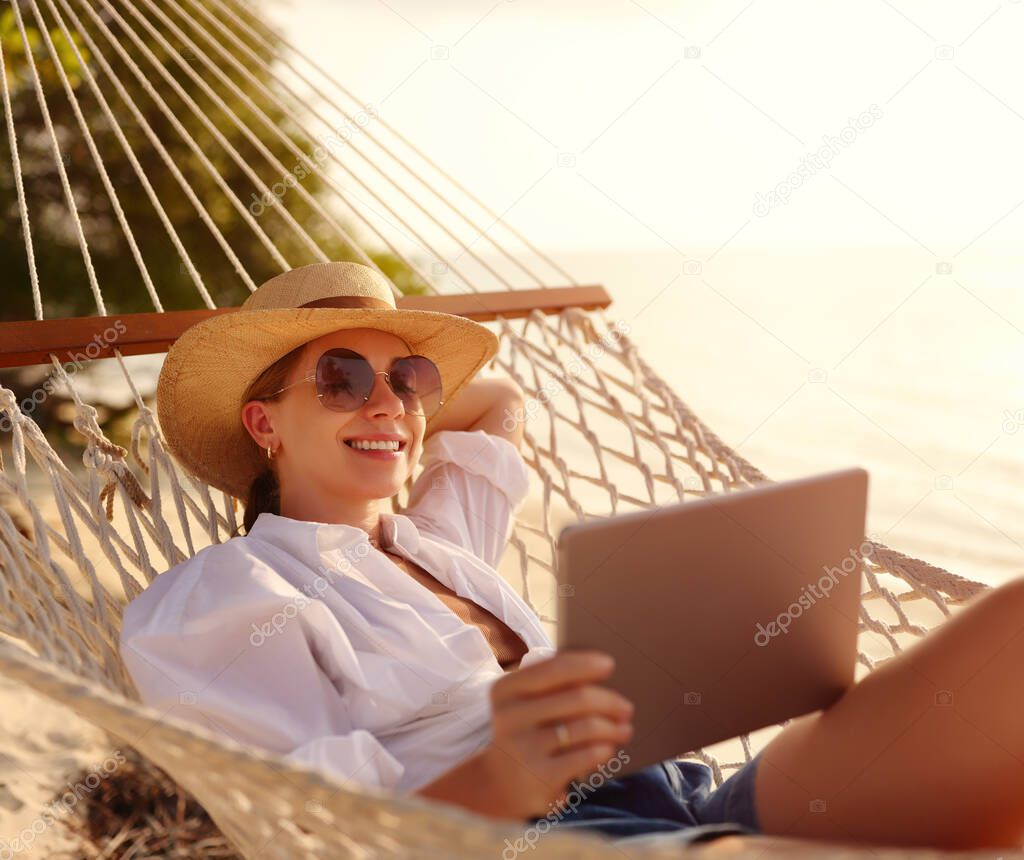 Young happy successful woman, smiling female freelancer in straw hat and sunglasses using digital tablet while lying and relaxing in the hammock on tropical sandy beach on hot sunny day. Distance work