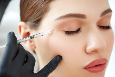 Güzellik kliniğinde cilt bakımı sırasında genç bayan hastaya iğne yapan eldivenli isimsiz bir kozmetik uzmanı.