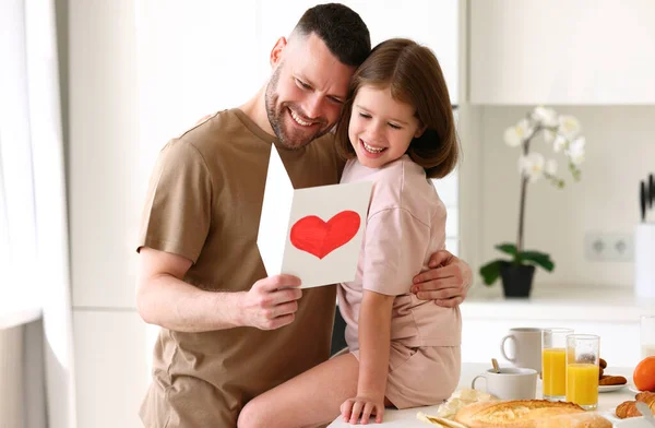 Little child daughter giving daddy greeting postcard with drawn red heart and smiling, celebrating family holiday together while having breakfast in kitchen at home. Fathers day concept
