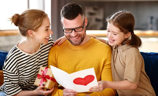 For best husband and dad. Happy family mother and little daughter congratulating father with holiday, giving him presents handmade postcard and wrapped gift box while sitting together on sofa at home