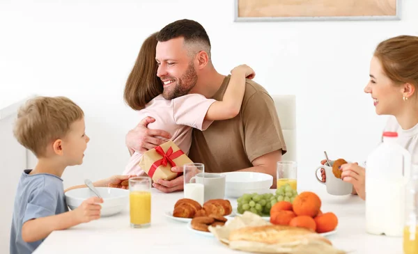 Best dad ever. Cute happy little girl daughter hugging with daddy, giving him wrapped gift box for Fathers day or birthday while eating breakfast with family in kitchen at home. Family holidays concept