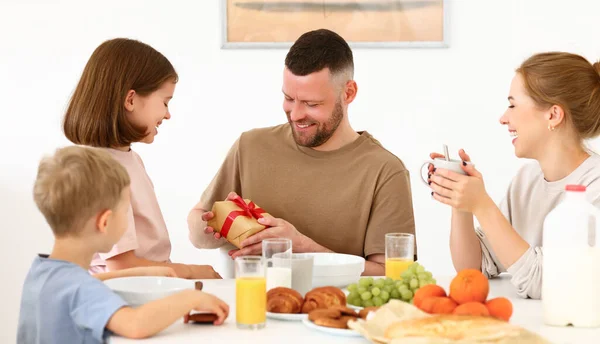 Best dad ever. Happy daddy holds in his hands a wrapped gift box given by cute children for the holiday  Fathers day or birthday while eating breakfast with family in kitchen at home. Family holidays concept