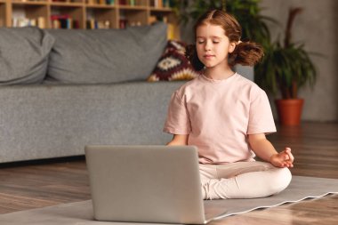 Çocuklar için yoga. Sakin, rahat giyinen, gözleri kapalı bir kız. Evde yoga dersine girerken dizüstü bilgisayarın önünde poz veriyor.