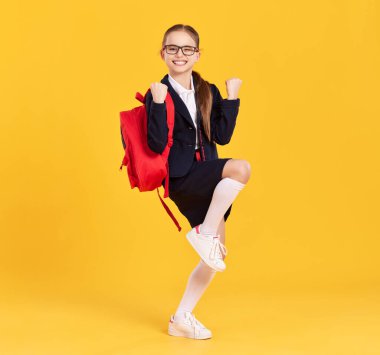 Üniformalı, gözlüklü, yumruklarını havada tutan ve sarı arka plandaki başarısını kutlayan zeki kız öğrenci.