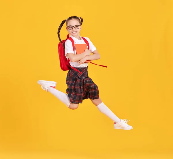 Full body of happy preteen schoolgirl in black uniform and glasses with backpack jumping high against yellow background