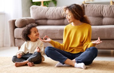 Çocuklu yoga. Genç Afro-Amerikan aile annesi ve küçük oğlu oturma odasındaki kanepenin altında oturmuş meditasyon yaparken gülümsüyor ve evde birlikte vakit geçiriyorlar.