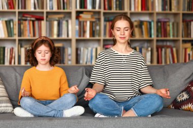 Genç anne ve kız evdeki oturma odasındaki kanepede birlikte otururken kapalı gözlerle meditasyon yapıyorlar. Covid19 karantinasında yoga yapan sakin bir aile.
