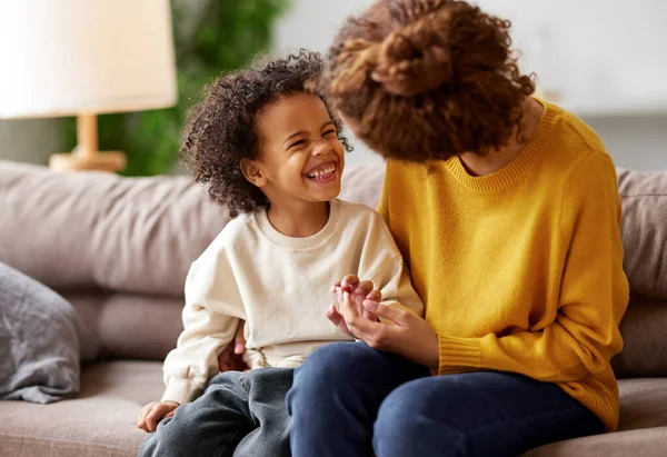 I love you, mom. Happy african american child son holding mothers hand, looking at her and smiling while sitting together on sofa in living room. Young family mom and kid enjoying time together at home