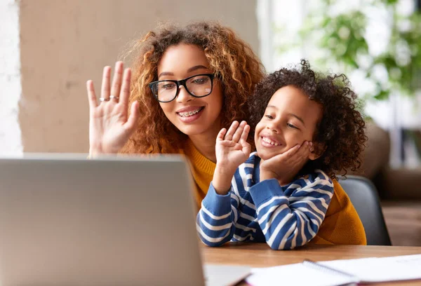 Happy afro american family, young positive mother and cute boy son having video call with relatives, father or grandparents, waving at webcam on laptop and smiling while sitting at desk at home