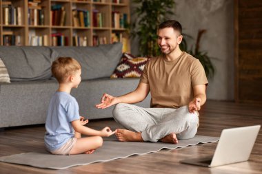 Gündelik giyinen genç, yakışıklı bir baba, oğlana Lotus 'ta birlikte oturma odasında otururken nasıl meditasyon yapılacağını öğretirken mutlu bir aile, dizüstü bilgisayarla yoga egzersizleri yapıyor.