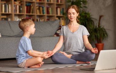 Bilgisayarla yoga yapan bir aile. Genç kadın anne ve küçük çocuk günlük giysiler giyip birlikte meditasyon yaparken oturma odasındaki dizüstü bilgisayarın önünde poz veriyorlar.