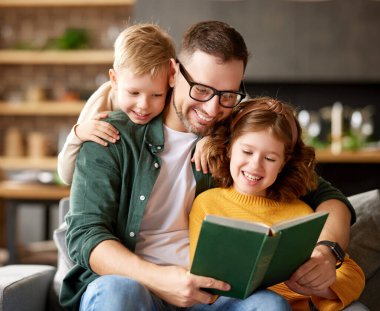 Gelmiş geçmiş en iyi baba. Genç, sevgi dolu bir baba, iki küçük tatlı çocukla kitap okuyor. Kanepede birlikte rahatlarken, mutlu bir aile kucaklaşıyor ve gülümsüyor.