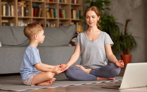 Family practicing yoga online with laptop. Young  woman mother and little child son wearing casual clothes meditating together while sitting in lotus pose on floor in front of laptop in living room