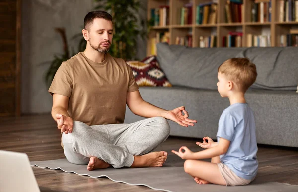 Family practicing yoga online with laptop. Young  man father and little child son wearing casual clothes meditating together while sitting in lotus pose on floor in front of laptop in living room