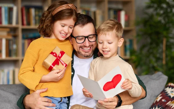 We love you, dad. Young happy father getting congratulations on Fathers Day from two excited kids at home, son and daughter, smiling children giving daddy handmade postcard and wrapped gift box