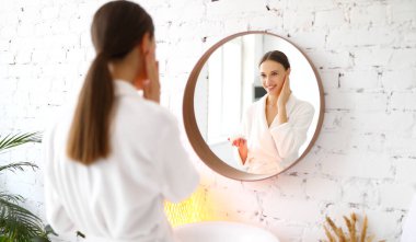 Bornozlu genç bir kadın yüzüne kozmetik krem sürüyor ve kaplıca güzellik seansı boyunca teninin yumuşaklığının tadını çıkarıyor.