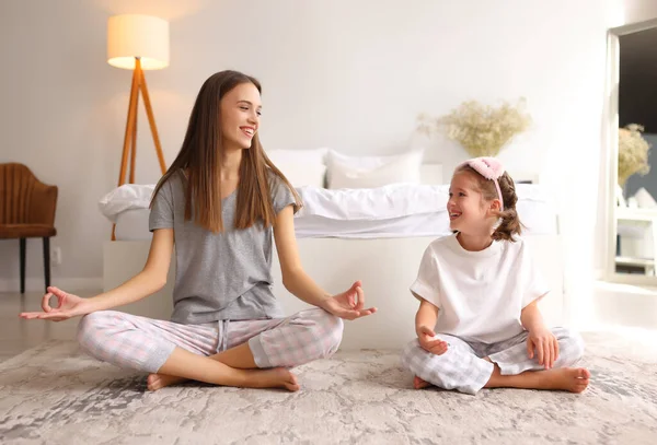 Full body family: young woman and girl sitting in Lotus pose and looking at each other with smile while meditating near bed at home