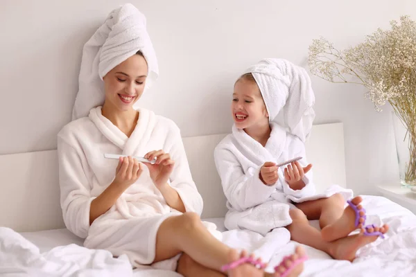 全身上下穿着浴衣和毛巾的年轻母亲和女儿在床上休息和在家里修指甲时 脸上挂着笑容 挂着钉子 — 图库照片
