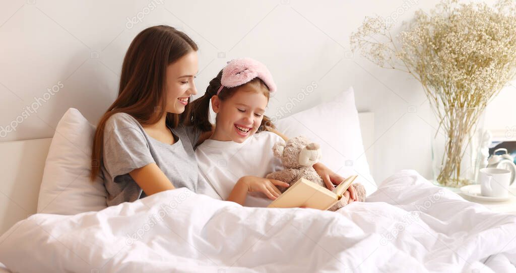 Cheerful mother hugging daughter while resting on bed and reading book together in weekend morning at home