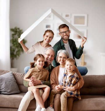 Büyük mutlu bir aile, büyük ebeveynler, anne, küçük çocukları olan baba yeni bir eve taşınmayı kutlayan oğul ve kız kağıt çatının altında kanepeye oturup kameraya gülümsüyorlar. İpotek kredisi kavramı