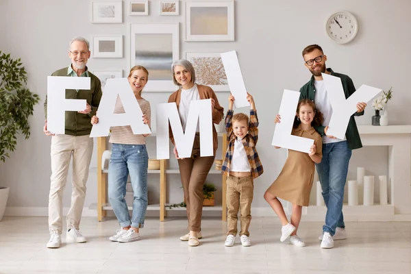 Big happy family in new house. Grandparents, mother and father with little kids holding word FAMILY and smiling at camera while standing in new modern appartment. Mortgage and real estate concept