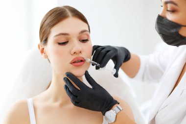 Genç bayan hasta, modern klinikte maske ve eldiven takan profesyonel kozmetik uzmanının dudaklarına enjekte ediliyor.